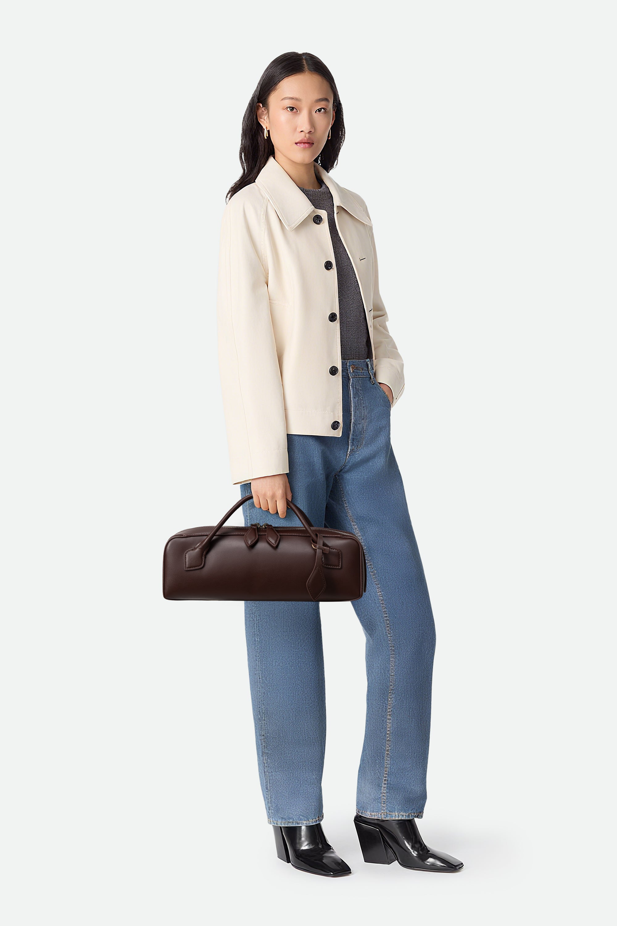 Woman holding a brown leather bag wearing a beige coat, gray shirt, blue jeans, and black shoes on a white background
