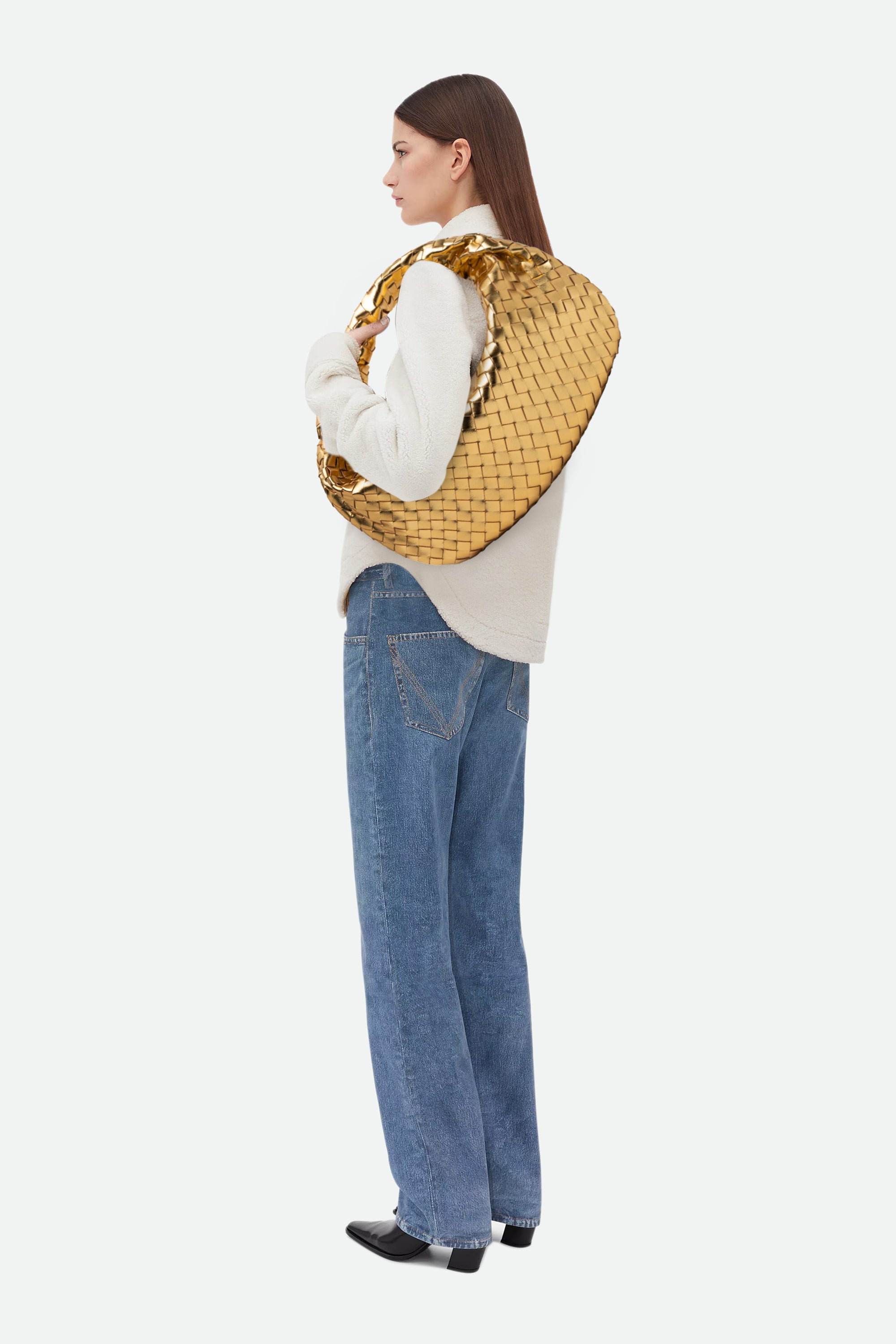 Woman wearing a light sweater and blue jeans carrying Large Knot Bag (Jodie) gold woven handbag on her shoulder.