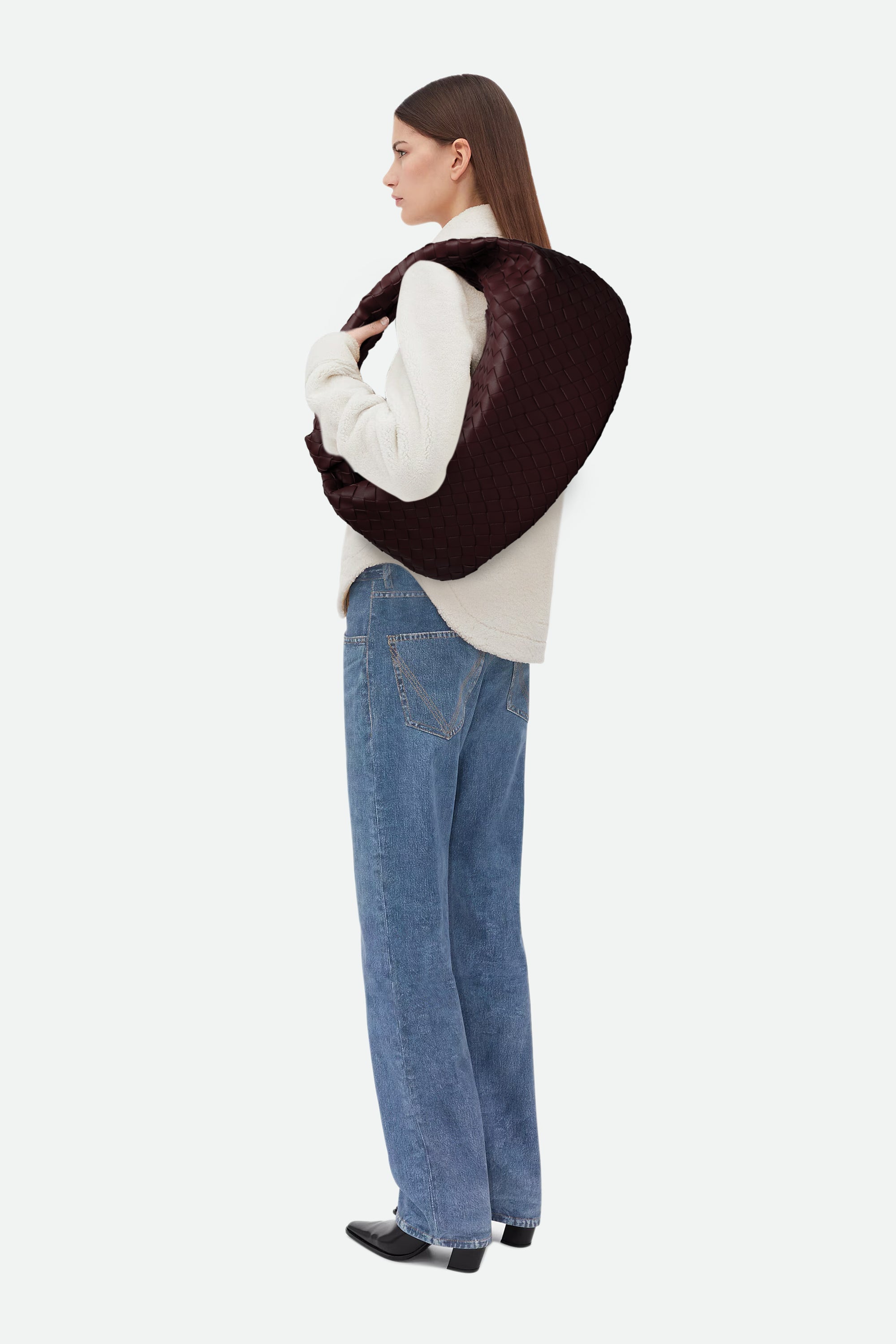 Woman wearing a white sweater and blue jeans carrying a wine red woven handbag on her shoulder