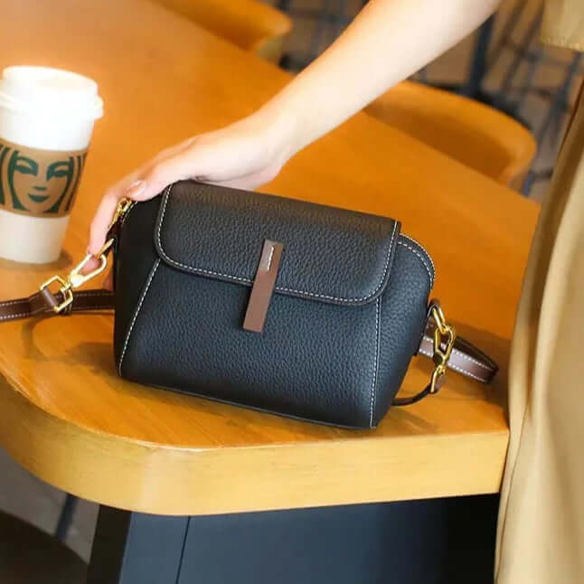 A black leather mini crossbody bag with a secure double-zip closure and adjustable straps, placed on a wooden surface with a cup of coffee and furniture in the background.