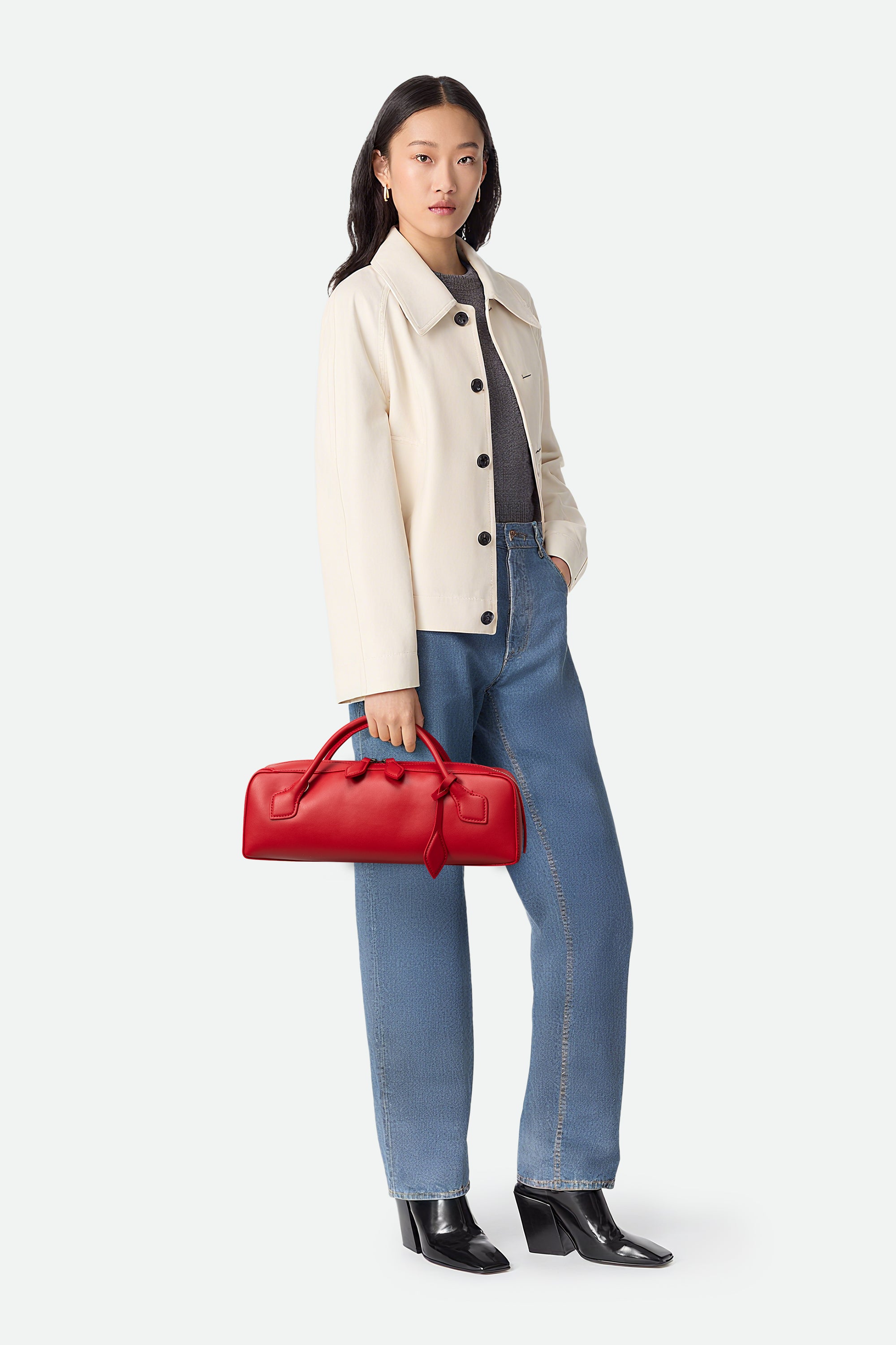 Woman holding a red handbag wearing a beige coat, gray sweater, blue jeans, and black shoes on a white background