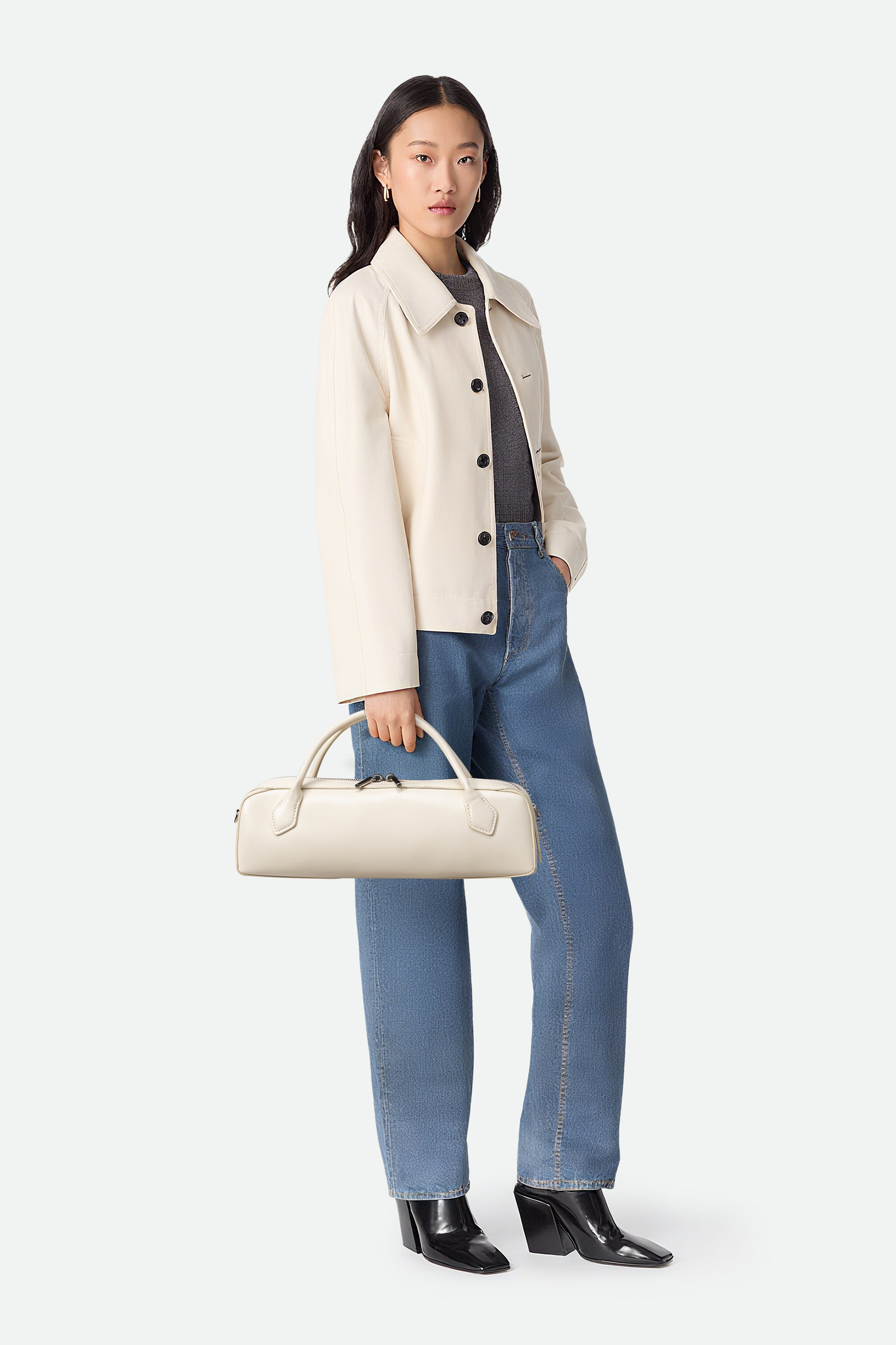 Woman wearing a beige coat, blue jeans, and holding a cream handbag on a white background