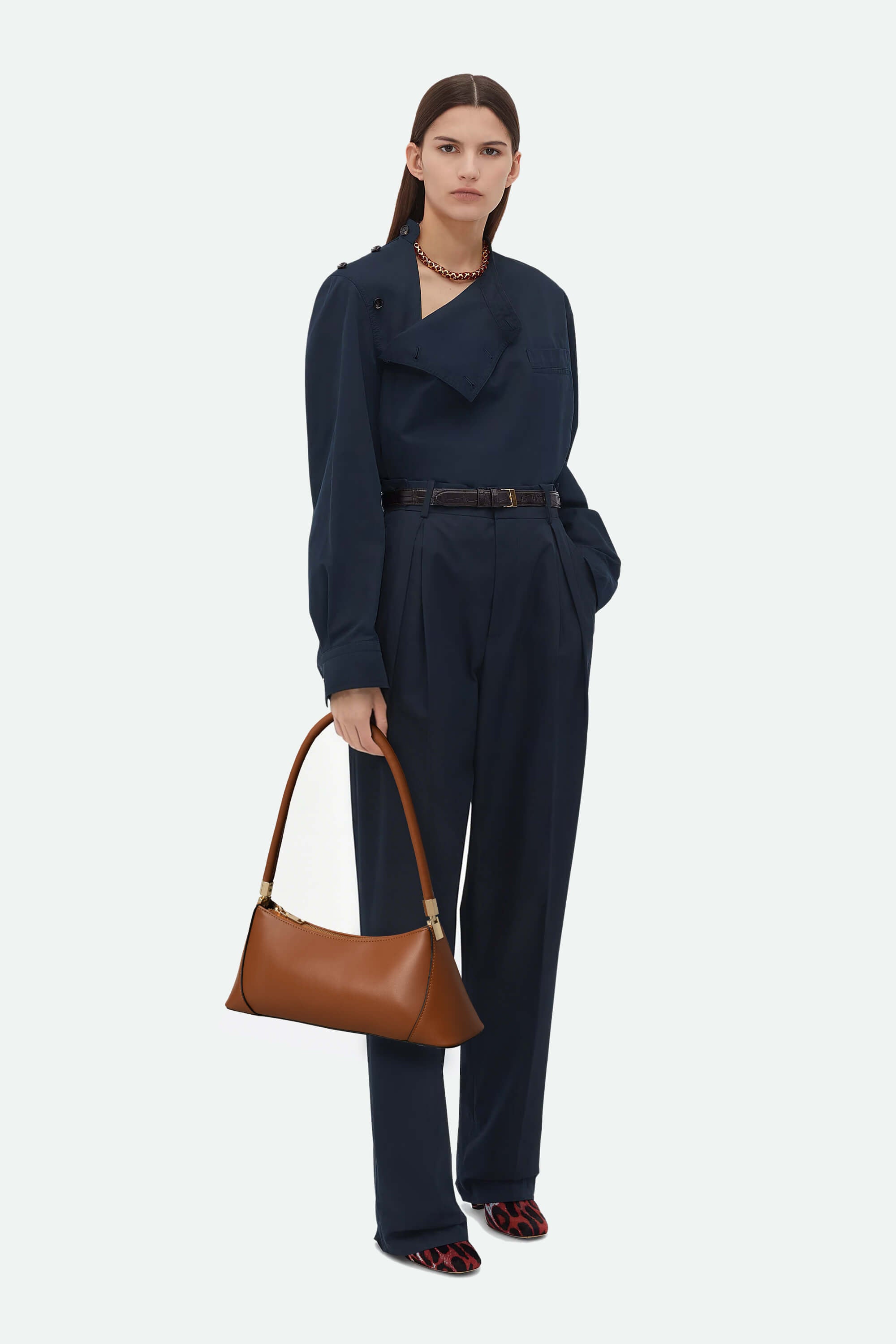 Woman wearing a navy blue jumpsuit holding a brown handbag on a white background